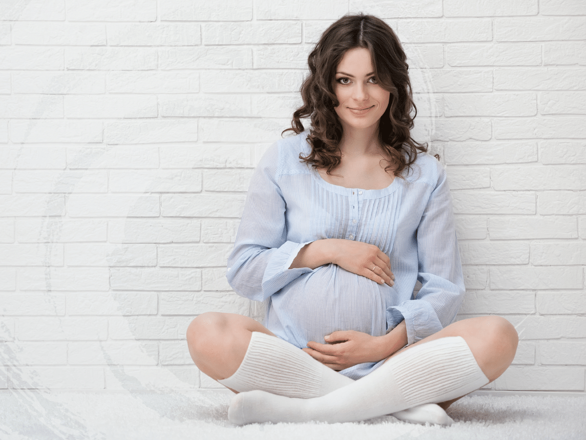 A pregnant woman sit crosslegged against a white brick wall, symbolizing TMS and pregnancy